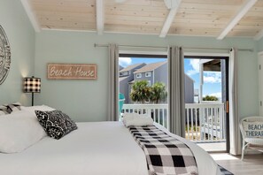 Een slaapkamer met een bed, een bordje met de tekst 'Beach House' en uitzicht op een balkon met een leuning en planten.