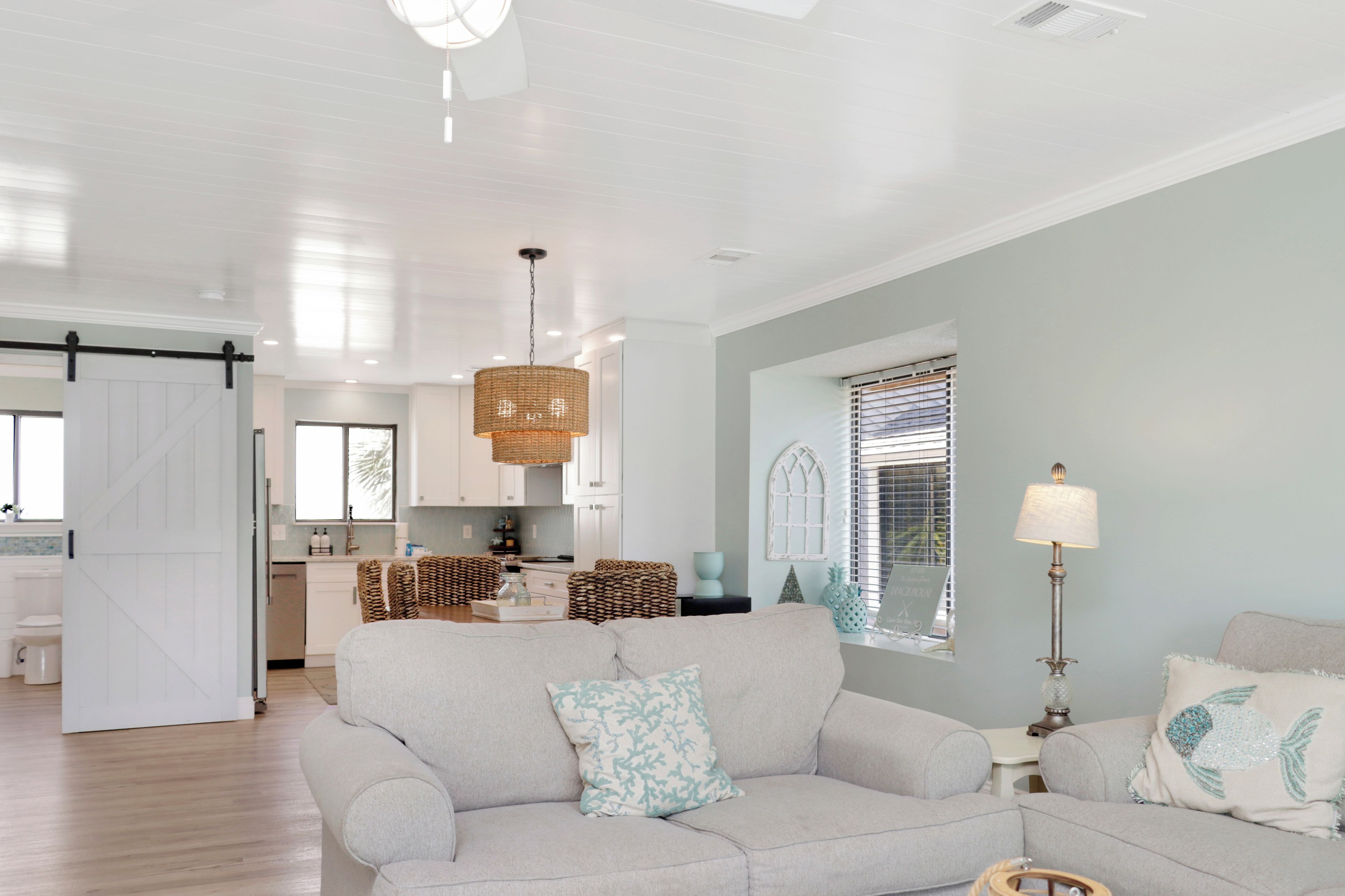 A spacious living room with a large grey sofa, a dining area with a kitchen in the background, and a bright ceiling with recessed lighting.