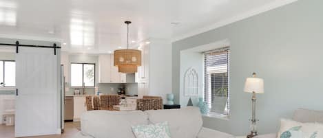 A spacious living room with a large grey sofa, a dining area with a kitchen in the background, and a bright ceiling with recessed lighting.