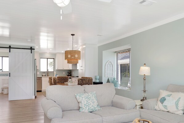 A spacious living room with a large grey sofa, a dining area with a kitchen in the background, and a bright ceiling with recessed lighting.