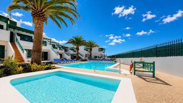 A pool area with clear blue water, lounge chairs, palm trees, and a building with balconies. - Chez Carmen • Pool View • Beach 900M • LVH (Puerto del Carmen)