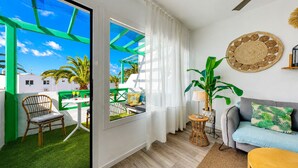 A room with a sliding glass door leading to a balcony, a sofa with a patterned cushion, a round woven wall decoration, and a small side table with a lamp. - Chez Carmen • Pool View • Beach 900M • LVH (Puerto del Carmen)
