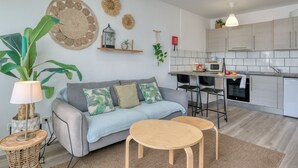 A modern kitchen with white cabinets, a dining area with a grey sofa and two wooden tables, and a living area with a large plant and decorative wall hangings. - Chez Carmen • Pool View • Beach 900M • LVH (Puerto del Carmen)