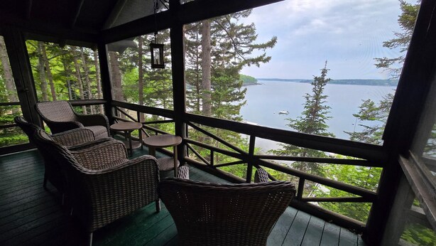 A wooden deck with wicker furniture and a view of a lake and forest. - Charming 4-Bedroom Linekin Log Cabin in Boothbay, ME with stunning waterfront views. (Boothbay)
