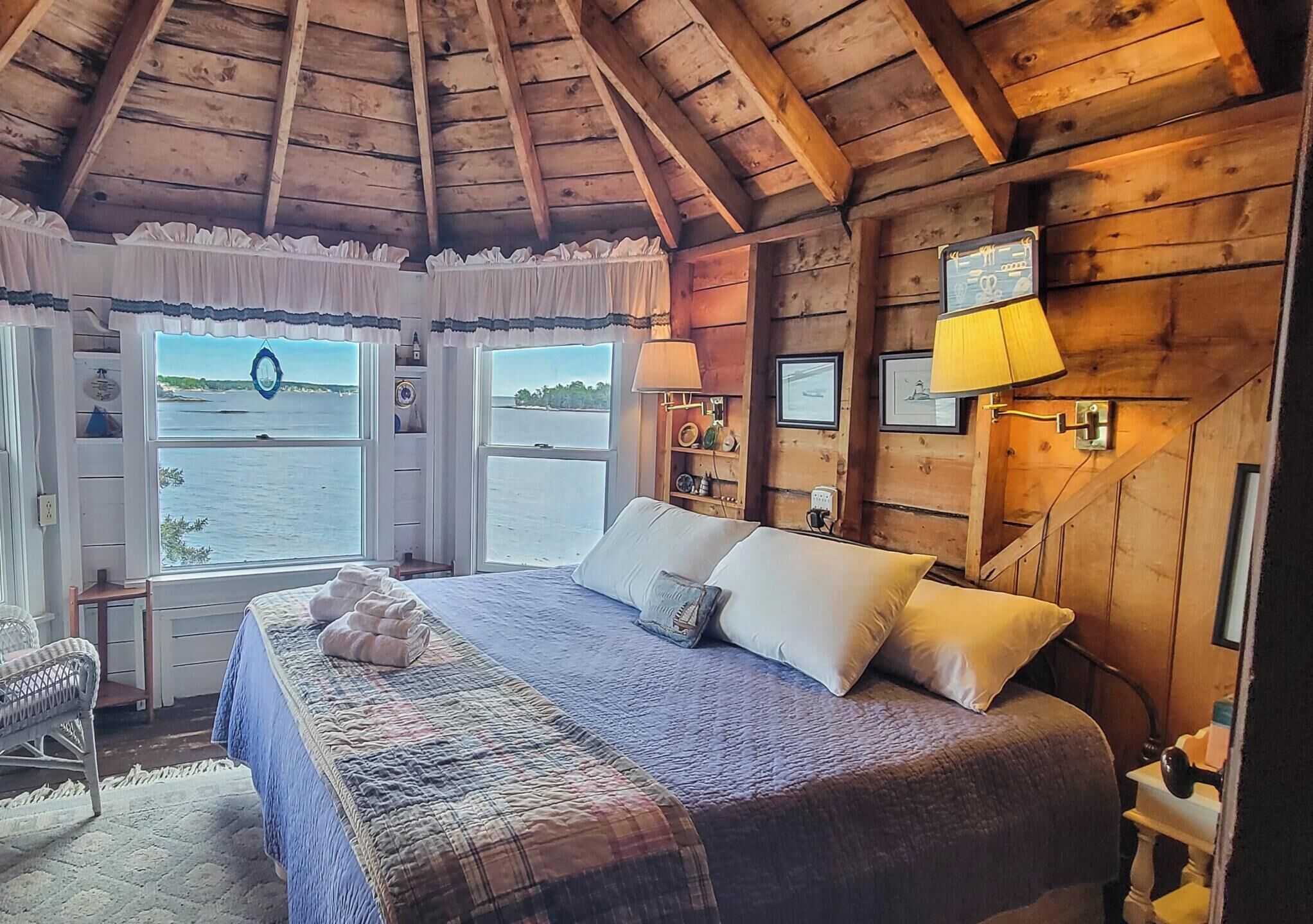 A cozy cabin bedroom with a large bed, a view of the water through windows, and wooden walls.