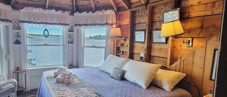 A cozy cabin bedroom with a large bed, a view of the water through windows, and wooden walls.