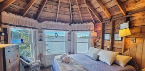 A wooden cabin bedroom with a large bed, two windows with a view of a lake, a wooden dresser, and a lamp. - Piffi Biff: Watch Whales & Lobster Boats from This Classic Oceanfront Gem! (Southport)