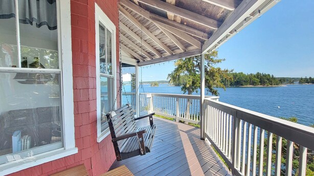 A wooden deck with a bench, a view of a lake, and a red building. - Experience lakeside luxury at Piffi Biff, a spacious 5-bedroom cabin on Southport, ME. (Southport)