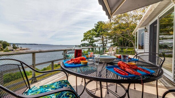 A round metal table with chairs, a lobster, and a pitcher of iced tea on a deck overlooking a body of water. - Experience coastal charm at Merwick Cottage in Georgetown, ME, with 3 bedrooms and waterfront views. (Georgetown)