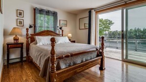 A bedroom with a wooden four-poster bed, a nightstand with a lamp, a large window with a view of a body of water, and a framed picture on the wall. - Experience coastal charm at Merwick Cottage in Georgetown, ME, with 3 bedrooms and waterfront views. (Georgetown)