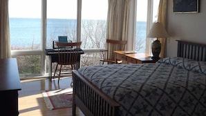 A bedroom with a bed, a desk, a chair, a lamp, and a view of the sea through large windows.