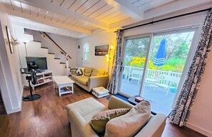 A cozy living room with a sofa, coffee table, and TV. There is a staircase leading to another level, and a balcony with a view of greenery.