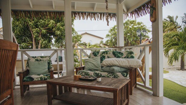 Um deck de madeira com um sofá e poltronas, uma mesa de centro e vista para um jardim com palmeiras.