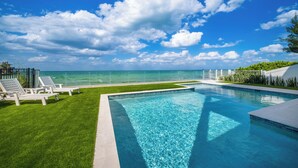 A swimming pool with a clear view of the ocean, surrounded by a well-maintained lawn and lounge chairs.