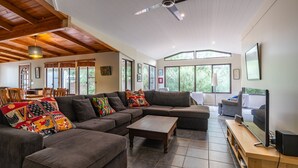A spacious living room with a large sectional sofa, a wooden coffee table, and a television on a wooden stand. There are large windows providing natural light and views of greenery outside.