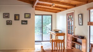 A dining area with a wooden table and chairs, a bookshelf, and a large window with a view of a deck and trees.