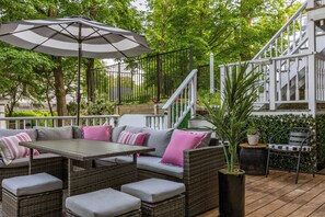 A patio area with wicker furniture, a table, and an umbrella. There is a staircase with white railings and a green hedge in the background. - Hot Tub + Walk to Lakeside Dining: Meredith Home! (Meredith)