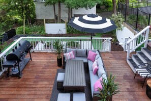 A wooden deck with a gray sofa, a grill, and a striped umbrella. - Hot Tub + Walk to Lakeside Dining: Meredith Home! (Meredith)