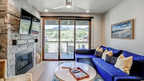 A modern living room with a stone fireplace, a blue sofa, and a large sliding glass door leading to a balcony with a mountain view. - Blue River Flats Retreat: Stunning Getaway With Amazing Views (Silverthorne)