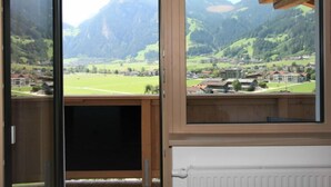 A room with a large window offering a view of a mountainous landscape and a village below. - Panoramablick by Interhome (Mayrhofen)