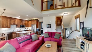 Amplia sala de estar con un sofá rojo, una mesa de centro de madera y un televisor sobre un mueble de madera. Se ve una cocina con armarios de madera y una zona de comedor con mesa y sillas.