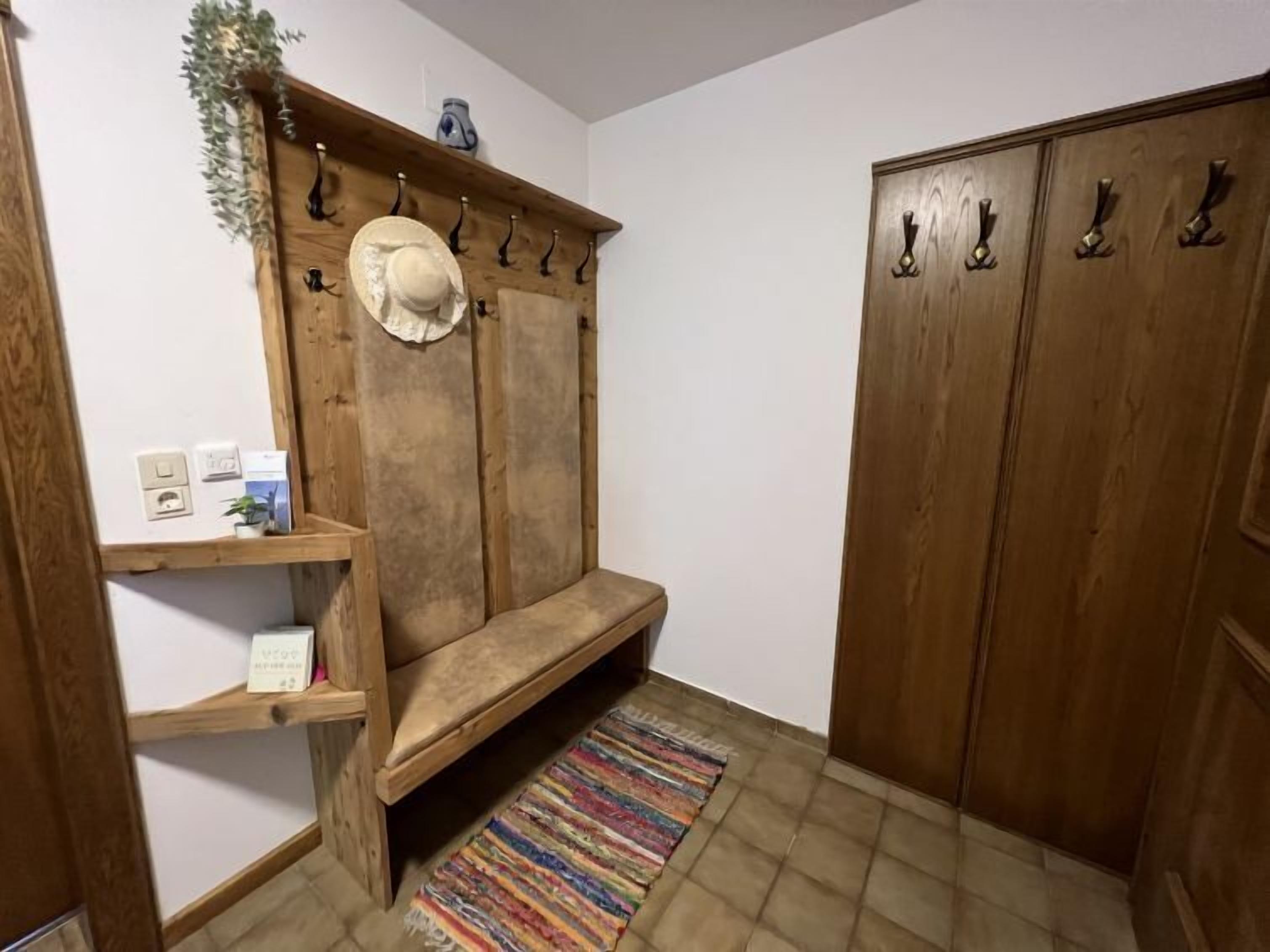 Eine hölzerne Bank mit einem Hutfächer darüber, ein kleines Regal mit einer Pflanze und ein farbenfroher Streifen Teppich auf dem Boden.