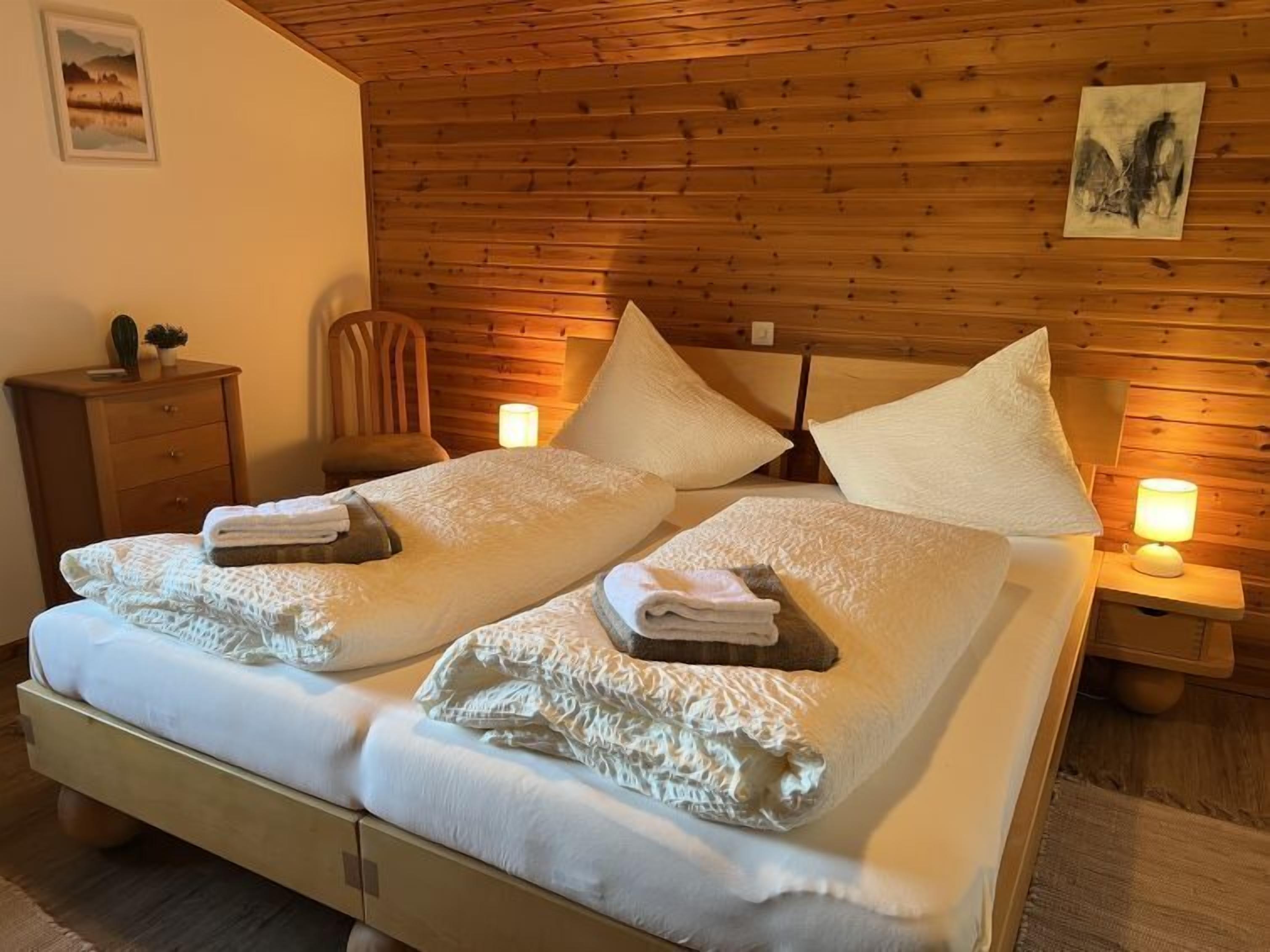 A double bed with white linens and pillows, a wooden headboard, a nightstand with a lamp, and a small wooden dresser.