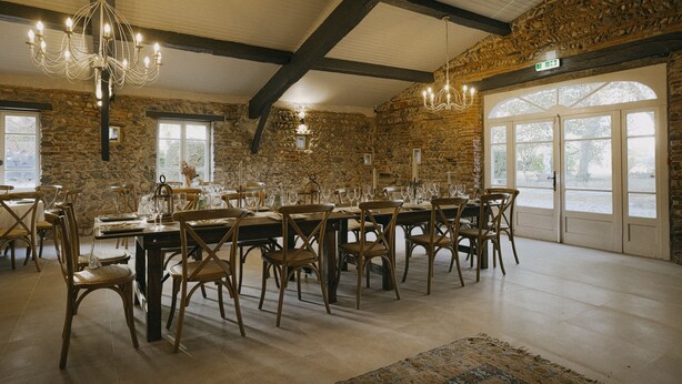 Un espace repas aux murs de pierre, avec des poutres en bois et des lustres. Les tables sont dressées avec des chaises et de la verrerie.