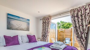 A bedroom with a bed, purple bedding, a painting of a beach, and a view of a wooden fence and greenery outside. - Charming Chalet A Stone's Throw From The Beach! - Pass the Keys (Chichester)