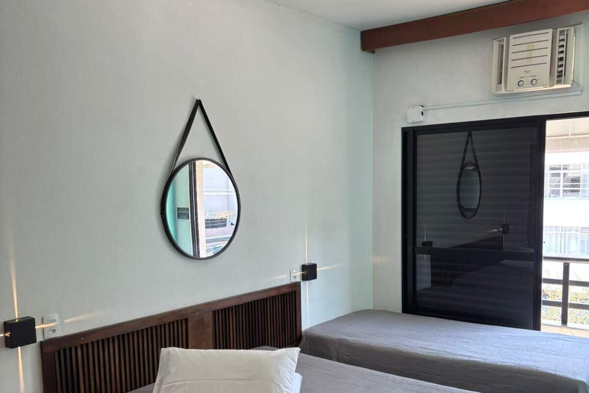 Habitación de hotel con dos camas, un espejo montado en la pared, un equipo de aire acondicionado y una ventana con persianas.