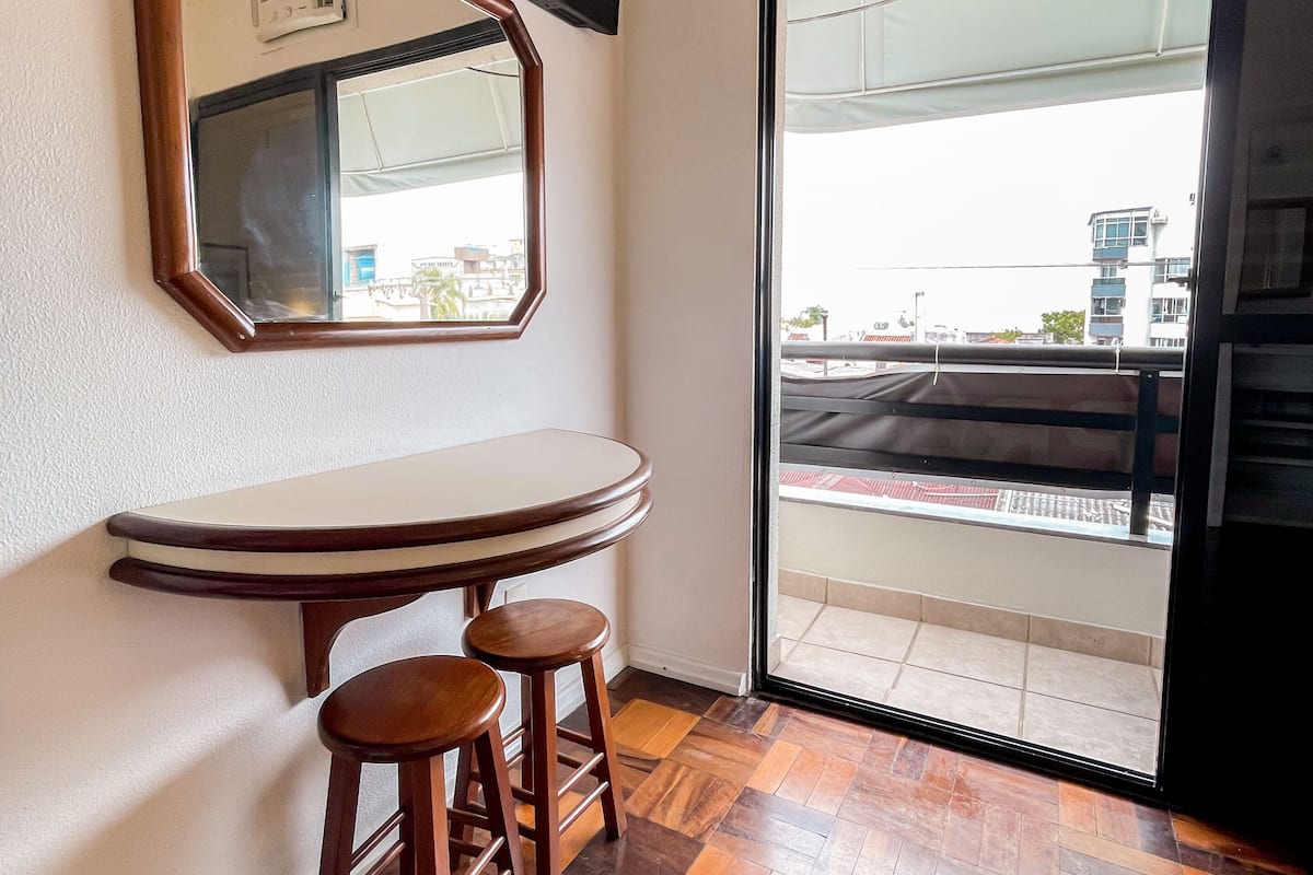 Una habitación con suelo de madera en patrón espiga, una mesa redonda con dos taburetes, un televisor montado en la pared y un ventanal corredero que da a un balcón exterior.