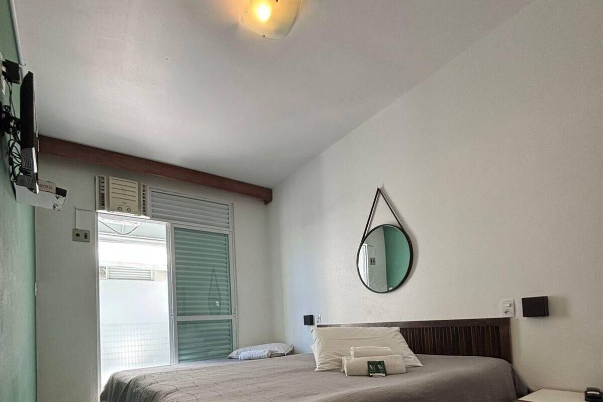 Un dormitorio con cama, mesita de noche, espejo, ventana con persianas y una lámpara en el techo.