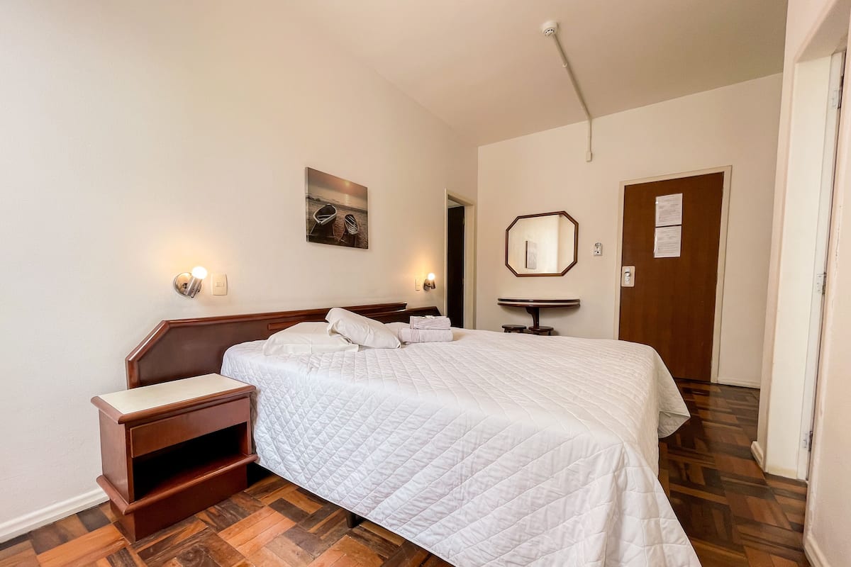 Habitación de hotel con suelo de madera, cama con ropa blanca, mesita de noche, espejo montado en la pared y pequeña mesa con espejo redondo.