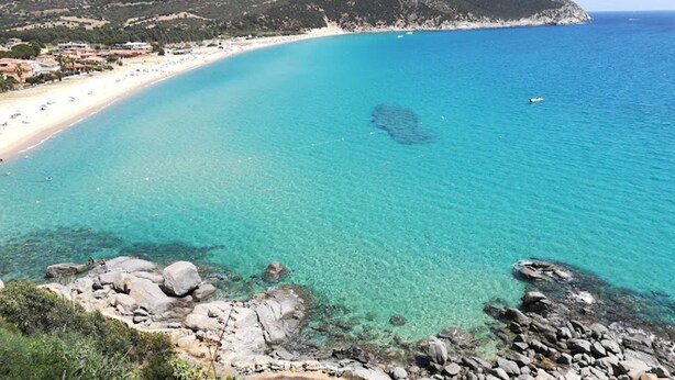 Un paysage côtier caractérisé par une eau turquoise et limpide, des rivages rocheux et une plage éloignée, parsemée de bâtiments.