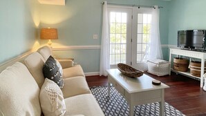 A living room with a white sofa, a ceiling fan, a television on a stand, and a window with curtains.