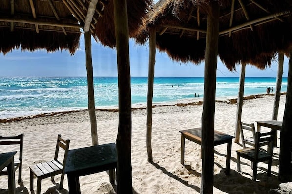 A beach with thatched-roof structures, wooden tables, and chairs.