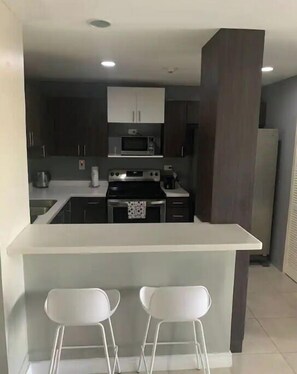 Una cucina moderna con mobili scuri, un piano di lavoro bianco e un forno a microonde integrato. Ci sono due sgabelli da bar bianchi al bancone.