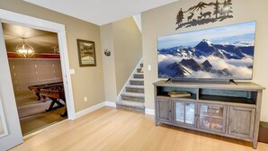 Sala de estar con piso de madera, un televisor montado en un mueble bajo, una escalera y una puerta que da a otra habitación con mesa de fútbol.