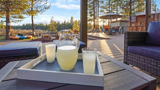 A wooden deck with outdoor seating, a glass pitcher, and two glasses of a beverage on a tray. - Riverfront Cabin w/ Trails, Hot Tub, Wood Stove, AC & River Views + dogs OK (Bend)