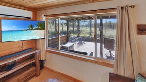 A room with a TV, wooden furniture, a large window with a view of a deck, and a chair. - Riverfront Cabin w/ Trails, Hot Tub, Wood Stove, AC & River Views + dogs OK (Bend)