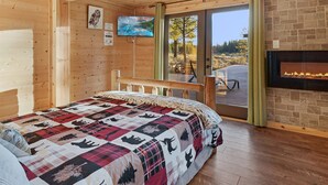 A wooden cabin bedroom with a large bed, a fireplace, and a scenic outdoor view. - Riverfront Cabin w/ Trails, Hot Tub, Wood Stove, AC & River Views + dogs OK (Bend)