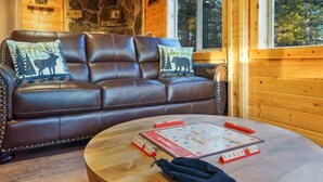 A brown leather sofa with two decorative pillows, a round wooden coffee table with a board game, and a stone wall in the background. - Riverfront Cabin w/ Trails, Hot Tub, Wood Stove, AC & River Views + dogs OK (Bend)
