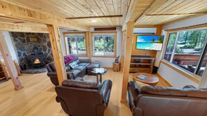 A cozy cabin interior with a stone fireplace, leather sofas, a television, and large windows offering a view of the forest. - Riverfront Cabin w/ Trails, Hot Tub, Wood Stove, AC & River Views + dogs OK (Bend)