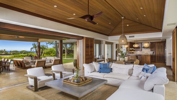 A spacious living room with a large sofa, coffee table, and dining area. There is a view of the outdoors and a kitchen area visible through an open door. - Elevated 4BR Kahikole Estate 218 at Hualalai – Indoor-Outdoor Living & Views (Kailua Kona)