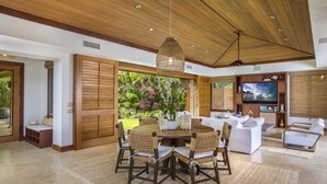 A spacious living area with a wooden ceiling, a dining table with woven chairs, a sofa set, and a television. - Elevated 4BR Kahikole Estate 218 at Hualalai – Indoor-Outdoor Living & Views (Kailua Kona)