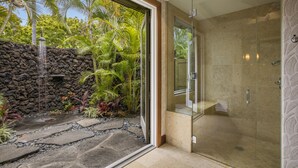 A modern bathroom with a glass shower enclosure, a stone wall, and tropical plants. - Elevated 4BR Kahikole Estate 218 at Hualalai – Indoor-Outdoor Living & Views (Kailua Kona)