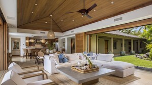 A spacious living area with a large white sofa, a coffee table, and a dining area with a kitchen in the background. - Elevated 4BR Kahikole Estate 218 at Hualalai – Indoor-Outdoor Living & Views (Kailua Kona)
