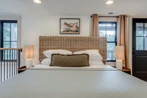 A bedroom with a large bed, a wicker headboard, two bedside lamps, a framed picture on the wall, and a window with curtains.