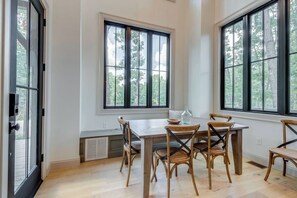 A dining area with a wooden table and chairs, large windows with a view of trees, and a door with a keypad lock.