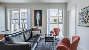 Ein modernes Wohnzimmer mit einer schwarzen Ledersofa, einem kleinen schwarzen Tisch und einem rosa Sessel. Große Fenster mit weißen Vorhängen, ein schwarzes abstraktes Gemälde und ein farbenfrohes abstraktes Bild schmücken die Wand.